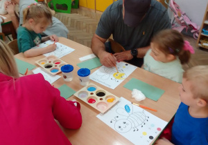 Julia, Tata Leny , Lena i Kamil przy stolikach wypełniają kolorowymi farbami plakatowymi kolorowankę pszczoły według podanego wzoru.