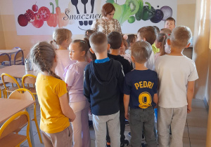Dzieci stoją przed Panią nauczycielką w stołówce szkolnej. W tle pomalowane na ścianie warzywa, sztućce, napis smacznego.