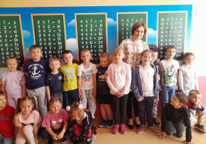 Dzieci wraz z Panią nauczycielką stoją przed powieszonymi na ścianie tablicami z tabliczką mnożenia.