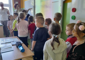 Przedszkolaki stoją w sali podczas lekcji religii. Mama Alicji pokazuje tablicę, obok stoi Pani prowadząca lekcję.