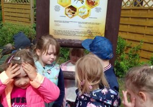 Dzieci stoją przy tablicy "Co nam daje pszczoła?" i słuchają informacji o produktach pozyskiwanych od pszczoły.
