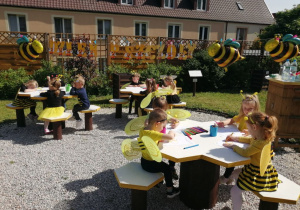 Dzieci siedzą przy stolikach i kolorują kolorowankę pszczółki.
