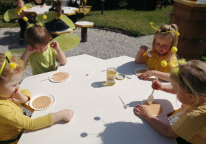 Grupka dzieci siedzi przy stoliku i smaruje wafelki miodem.