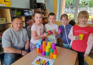 Mama Alicji i tata Krzysia oraz Wiktoria, Krzyś, Alicja i Alicja prezentują przy stole zbudowaną według kodu wieżę z kolorowych kubków.