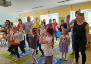 Dzieci razem z rodzicami tańczą na dywanie przy piosence "Matematyczna gimnastyka". Wszyscy podnoszą ręce do góry.