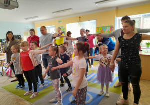 Dzieci razem z rodzicami podczas zabawy tanecznej na dywanie wyciągają ręce na boki.