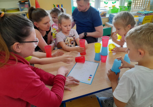 Dzieci razem z rodzicami siedzą przy stole i budują z kolorowych kubków wieżę według kodu.