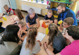 Dzieci przy dwóch stołach układają wspólnie z rodzicami wieże z kolorowych kubków według kodu.