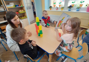 Oskar, Winicjusz i Anton siedzą przy stole razem z rodzicem - mamą Oskara. Na środku stołu znajduje się zbudowana przez nich wieża z kolorowych kubków.