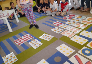 Wiktoria stoi na dywanie i liczy elementy na karcie. Na dywanie leżą karty z różną liczbą elementów oraz z cyframi. W tle rodzice siedzą na krzesłach, a dzieci na dywanie.