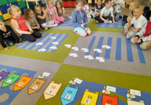 Przedszkolaki siedzą w półkolu. Na środku dywanu leżą odwrócone karty z pszczółkami i kolorowe ule z cyframi. Alicja odczytuje działanie matematyczne na wybranej przez siebie karcie.