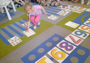 Natalia podczas gry "Wiosenne kodowanie" układa na dywanie kartę według kodu. Działania dziewczynki obserwują dzieci i rodzice.