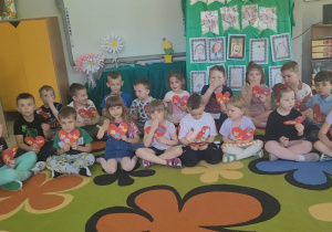 Dzieci z grupy "Biedronek" siedzą na dywanie i śpiewają piosenkę dla mam. W dłoniach przedszkolaki trzymają laurki w kształcie serduszek.