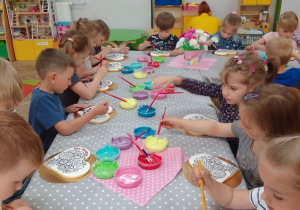 Dzieci z grupy "Motylków" przy stolikach ozdabiają pędzelkami i kolorowym lukrem ciastka w kształcie serca z czerwonym napisem Dla Mamy. Na stoliku są pojemniki z kolorowym lukrem, pędzle, kolorowe posypki.