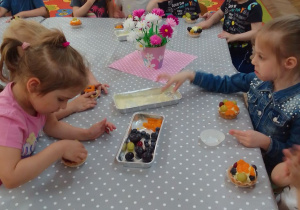"Motylki" dekorują kremowe babeczki owocami: winogronem i kawałkami pomarańczy. Na stole koszyk z różowymi i białymi stokrotkami z bibuły.