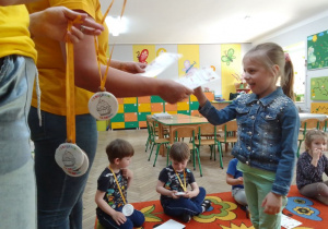 Zuzia odbiera z rąk Pani Emilii dyplom i jadalny medal z piernika. Pozostałe dzieci siedzą na dywanie.