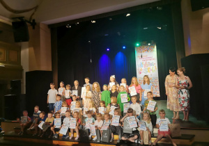 Na scenie CTMiT stoją i siedzą wszystkie dzieci, które wzięły udział i zostały nagrodzone w Festiwalu piosenki. Dzieci z uśmiechami na twarzy pozują do zdjęcia trzymając w rękach otrzymane dyplomy i nagrody.