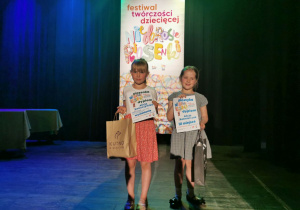 Ala Ś i Ala K. stoją na scenie CTMiT na tle plakatu Festiwal Twórczości Dziecięcej "Niedorosłe piosenki". Dziewczynki pozują do zdjęcia trzymając otrzymane dyplomy i nagrody.