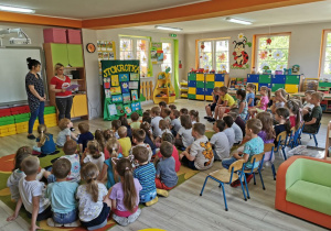 Dzieci z wszystkich grup przedszkolnych siedzą na dywanie i krzesełkach w sali. Przed tablicą stoją dwie panie, które ogłaszają wyniki konkursu.