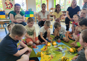 Dzieci z grupy Biedronek siedzą wokół maty do kodowania „Mapy skarbów” i programują pszczoły Bee Bot. W tle widać zgromadzonych rodziców