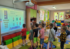 Przedszkolaki stoją ustawione w rzędzie przed tablicą multimedialną, na której wykonują quizy matematyczne. Działaniom dzieci przyglądają się rodzice