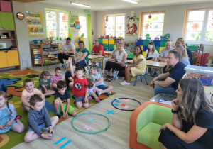 Dzieci siedzą na dywanie i wykonują działania matematyczne polegające na dodawaniu na konkretach. W tle widać zgromadzonych rodziców