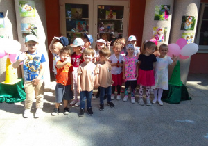 Grupa Motylków na tarasie pozuje do zdjęcia podczas uroczystości Dnia Dziecka. Z obu stron kolumny, na których są obrazki z postaciami dzieci oraz białe i różowe balony.