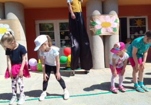 Zuzia, Julia, Amelia i koleżanka z grupy Słoneczek przechodzą po linie bokiem. Z tyłu szczudlarz, kolorowe balony a na kolumnach obrazy stokrotek wykonane przez dzieci podczas obchodów Dnia Stokrotki.