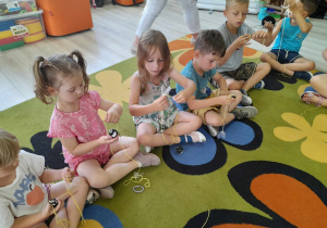 Kolejna grupka dzieci siedzi obok siebie na dywanie. Dzieci nawijają żółtą włóczkę ze skrzydełkami na szyszkę.