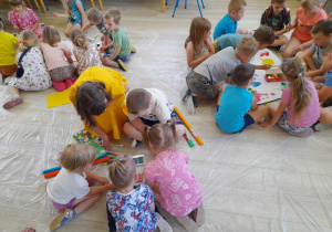 Dzieci siedzą na dywanie podzielone na trzy grupy i ozdabiają elementy motyli. Z dziećmi siedzi Pani prowadząca warsztaty.