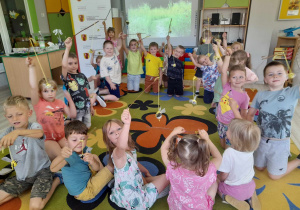 Dzieci siedzą na dywanie i prezentują przymocowane do kijków trzmiele. W tle tablica multimedialna, rolap.