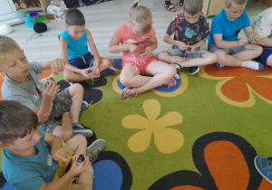 Kilkoro dzieci siedzi obok siebie na dywanie i wykonują trzmiela. Przedszkolaki nawijają żółtą włóczkę na szyszkę.