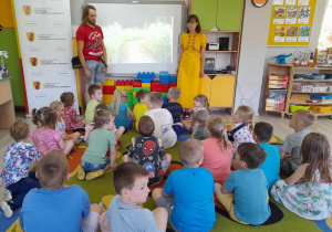 Dzieci siedzą na dywanie przed tablicą multimedialną. Przed dziećmi stoi Pani i Pan prowadzący zajęcia, a obok tablicy znajduje się rolap.