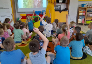 Dzieci siedzą na dywanie przed tablicą multimedialną. Pani prowadząca warsztaty rozmawia z dziećmi na temat owadów zapylających. Przedszkolaki zgłaszają się do odpowiedzi podnosząc ręce do góry.