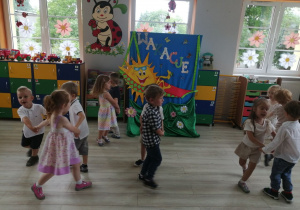 Dzieci tańczą w małych kółeczkach na tle dekoracji z okazji zakończenia roku szkolnego.