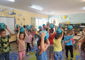 Dzieci z grupy "Słoneczek" spacerują po sali trzymając nad głowami niebieską piłkę.