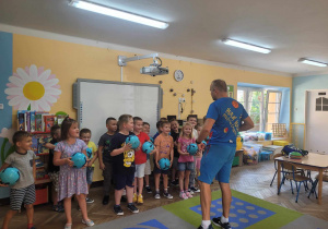 Przedszkolaki z grupy 'Słoneczek" stoją w sali przed tablicą multimedialną. Dzieci trzymają w dłoniach piłki. Przed nimi stoi instruktor Drużyny Kangura