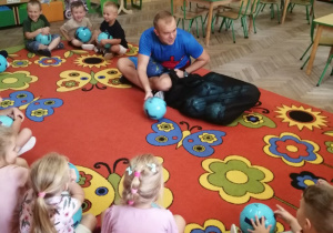 Dzieci siedzą w kole na dywanie, a pan instruktor rozdaje niebieskie piłki.