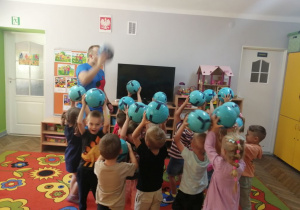 Dzieci stoją z piłkami uniesionymi do góry, obok grupki dzieci instruktor w niebieskim stroju.