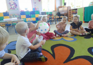 Grupa Biedronek na dywanie w kole, dzieci przekazują sobie balon w kropki przy dźwiękach muzyki.