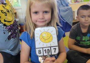 Lena pokazuje kartonik ze słoneczkiem, na którym przypięła klamerkę. Klamerka wskazuję cyfrę odpowiadającą liczbie piegów słoneczka.