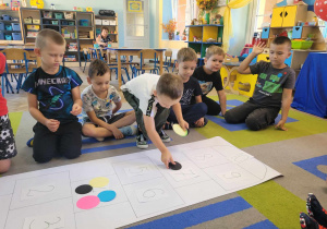Przedszkolaki siedzą na dywanie przed dużym kartonem, na którym układają kolorowe kropki.
