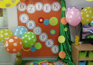 Pomarańczowa tablica, na której znajduje się napis "Dzień kropki" oraz kolorowe kropeczki. Na szafce stoją kolorowe balony w kropki oraz kolorowe tacki na których leżą papierowe kropeczki.
