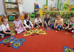 Dzieci siedzą na dywanie, trzymając kredki którymi rytmicznie uderzają o kolorowe kropki leżące przed nimi.