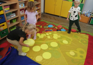 Grupka dzieci stoi na chuście animacyjnej i kładzie na niej żółte kropeczki.