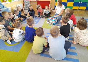 Dzieci siedzą na dywanie, a Pani ze szkoły językowej przedstawia dzieciom maskotkę - pluszowego misia.