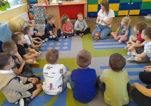 Dzieci siedzą na dywanie w kole razem z Panią ze szkoły językowej. Pani pokazuje przedszkolkom obrazek z książką, a przed nią leży maskotka - pluszowy miś.