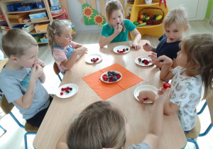 Dzieci przy stole degustują owoce: śliwki, maliny i jeżyny przyniesione ze sklepu.