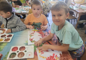 Eryk, Franio i Tymek siedzą przy stolikach w trakcie malowania farbami jesiennych liści.