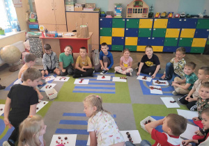 Dzieci siedzą w kole na dywanie. Przed nimi widać rozłożone białe kartony, na których przedszkolaki układają obrazki z materiału przyrodniczego.